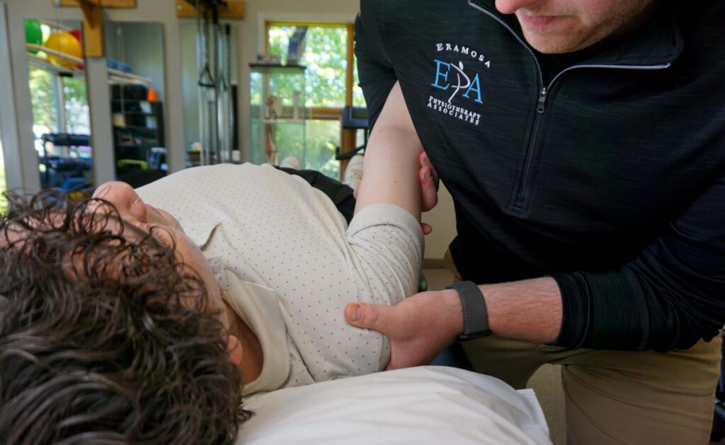 A physiotherapist in an Eramosa Physiotherapy fleece performing hands-on shoulder and arm treatment with a patient lying on a treatment table